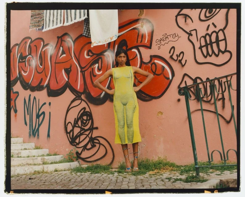 Woman poses in a yellow dress with graphic prints on a street in front of a pink wall with graffiti on a sunny day.