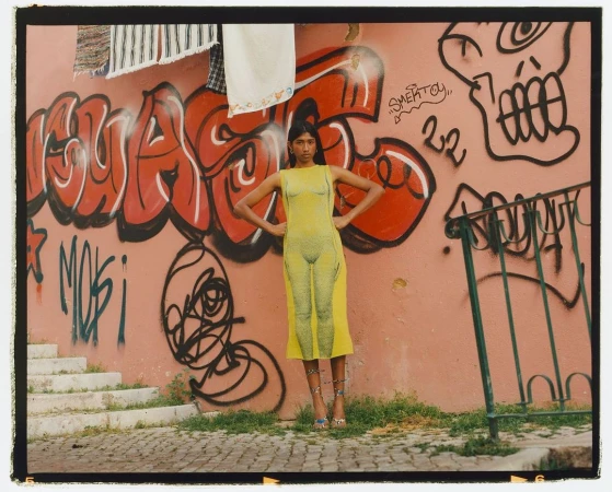 Woman poses in a yellow dress with graphic prints on a street in front of a pink wall with graffiti on a sunny day.