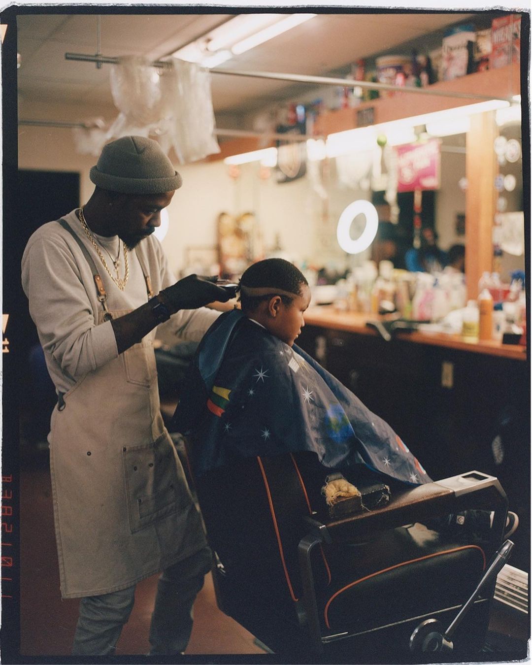 A barber in a grey beanie gives a young child a haircut in a vintage-style barber's chair inside a brightly lit barbershop.