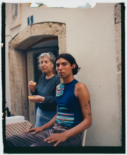 A young adult sits outside next to an older woman holding orange slices, photographed with natural light in an urban setting.