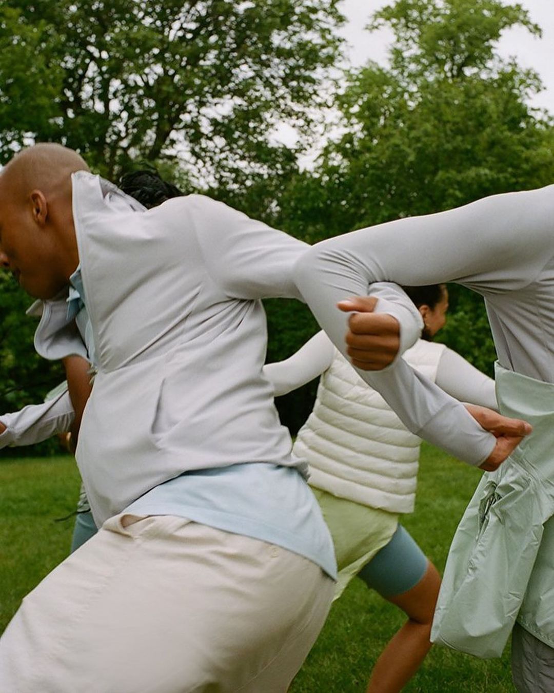 Group of people in athletic clothing moving on green lawn, surrounded by trees in an outdoor setting under an overcast sky.