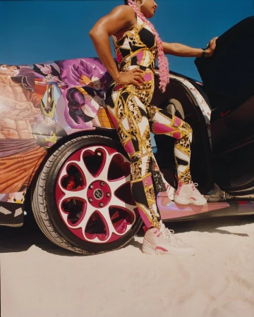 Adult female poses next to an automobile with customized rims and illustration, wearing patterned jumpsuit, jewelry, and light pink sneakers outside.