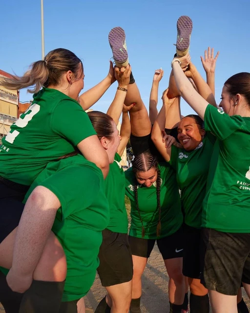 Female athletes celebrate teammate by lifting her up in air. Group wears green shirts, black shorts, outdoors, blue sky overhead. Sports.