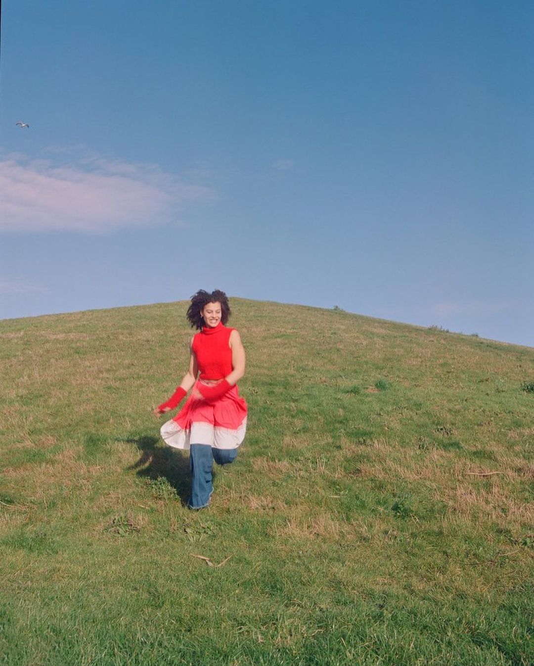 Woman with curly hair wearing a red top, running down a grassy hillside in a bright outdoor daytime setting under a clear blue sky.