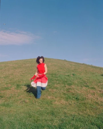 Woman with curly hair wearing a red top, running down a grassy hillside in a bright outdoor daytime setting under a clear blue sky.