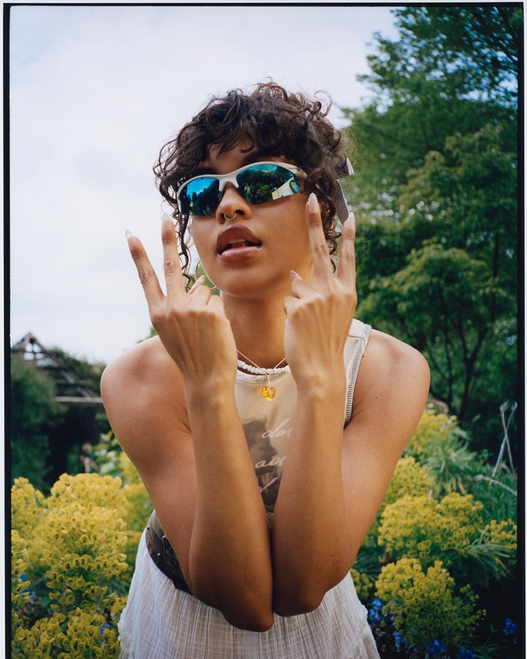 Young woman in sunglasses poses with a peace sign in a sunlit outdoor garden with yellow flowers and green foliage. Casual fashion shoot.