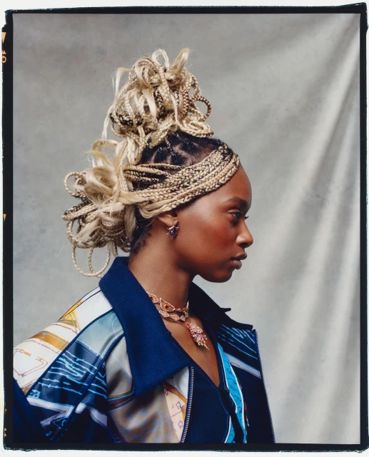 Woman in profile wearing an elaborate braided hairstyle with a printed jacket in a studio setting with soft, diffused lighting.