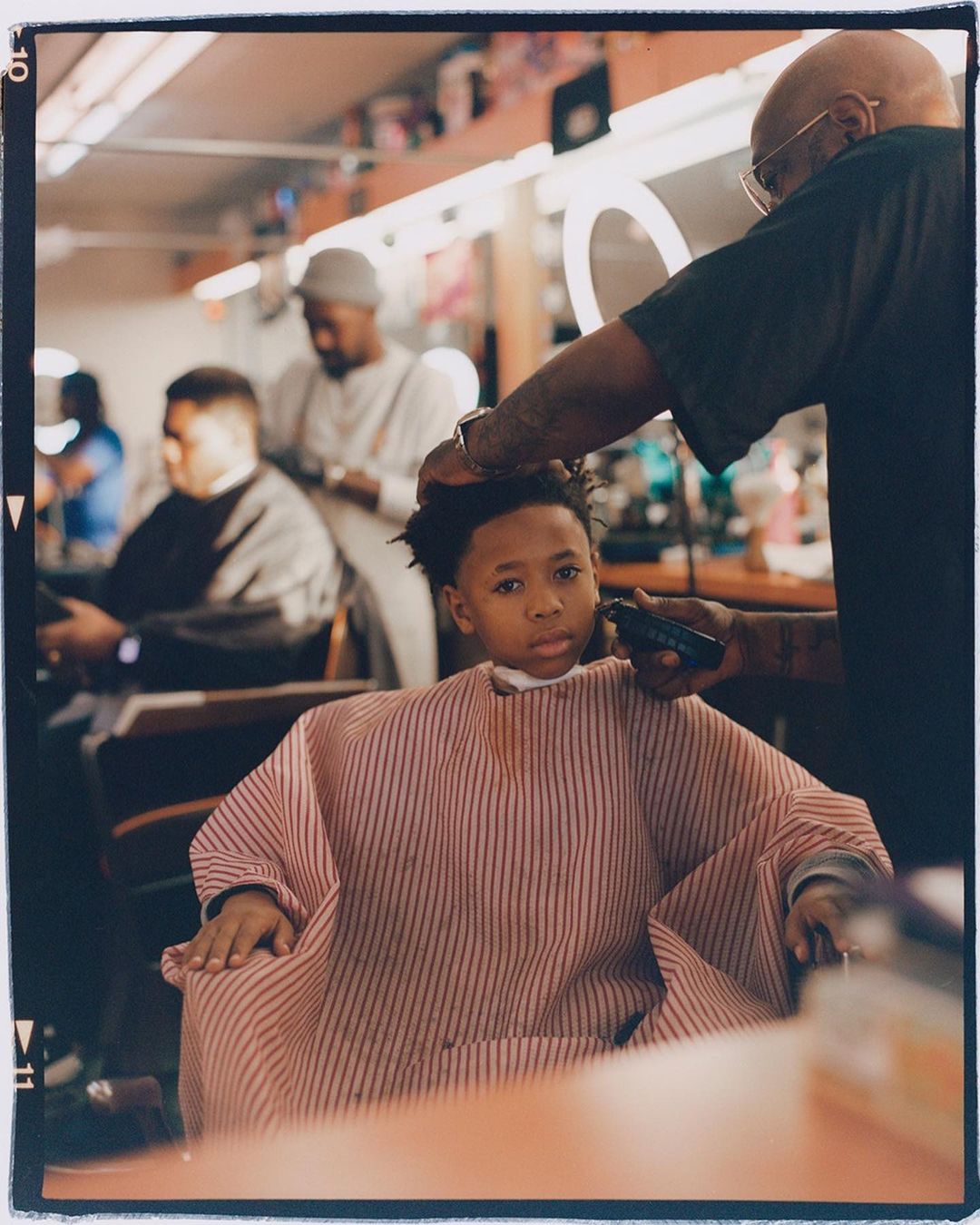 Young boy getting a haircut at a barber shop. A barber is using clippers to trim the boy's hair. Other customers are waiting in the background.