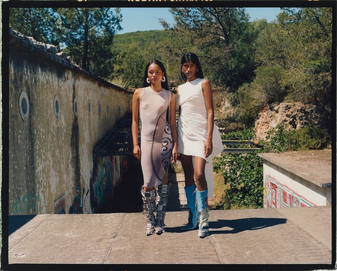 Two women pose outdoors in fashion dresses and embellished boots in front of an old building on a sunny day, showcasing style.
