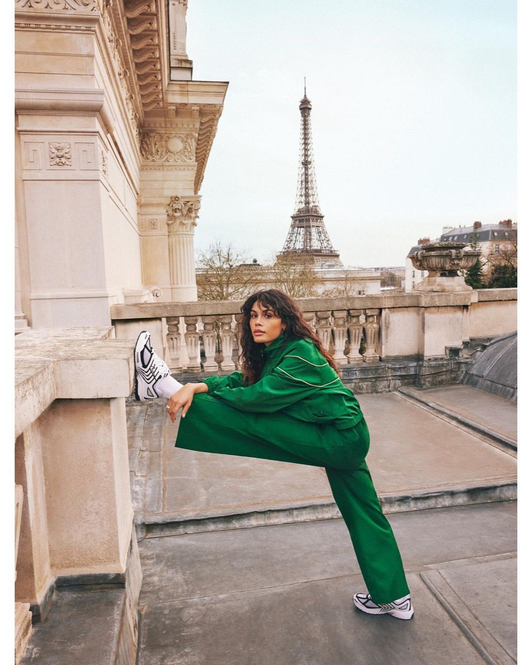 A woman in a green tracksuit stretches on a Parisian rooftop with the Eiffel Tower in the background on a cloudy day.