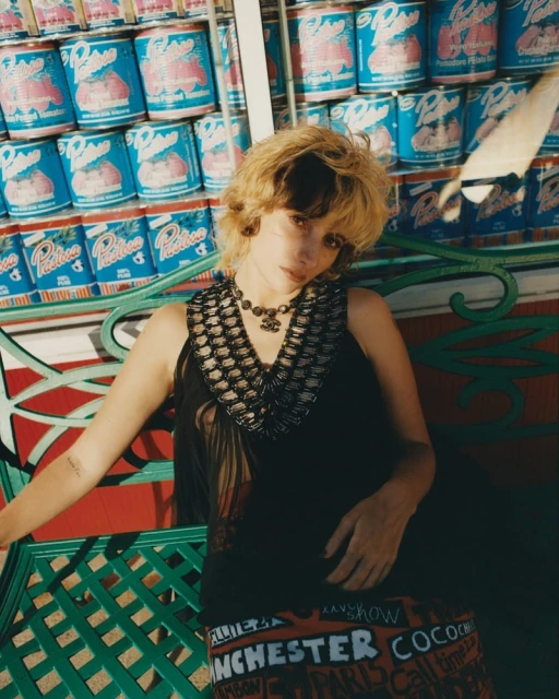 Stylish woman in black top and patterned skirt posing outdoors against a backdrop of stacked cans, accessorized with necklace and earrings.