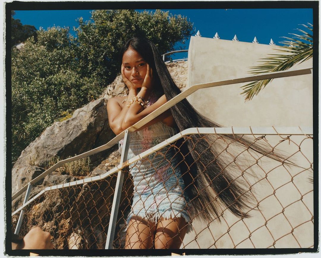 Young Asian woman poses outdoors against a railing with trees and a blue sky backdrop, showcasing her long dark hair and light dress.