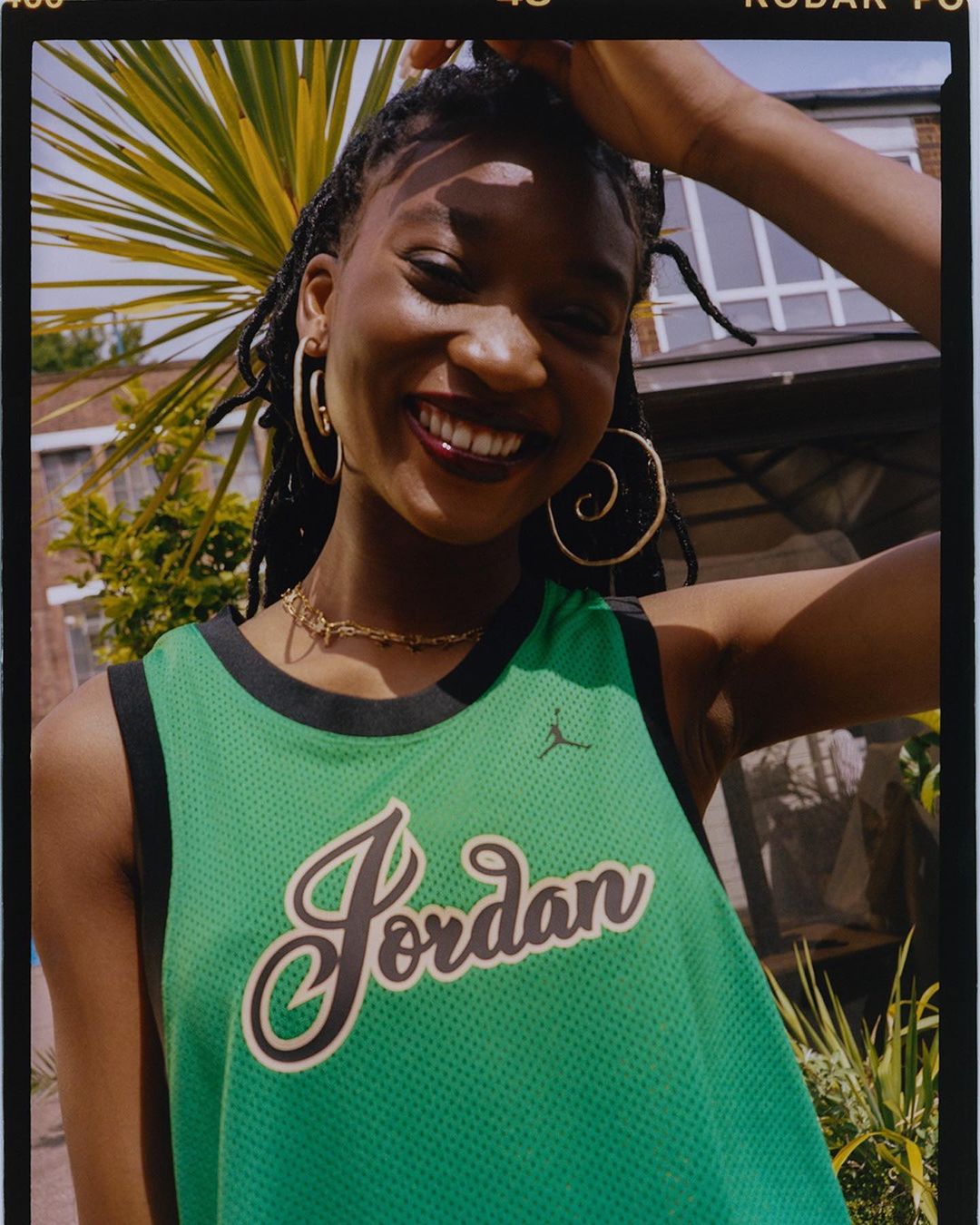 Smiling Black woman in a green Jordan jersey outside with palm trees. Close-up of a happy, fashionable young adult in natural light.