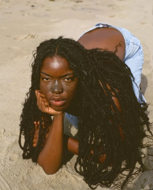 Portrait of a Black woman with dark hair, wearing denim, lying on a sandy beach. She is resting her head on her hand.