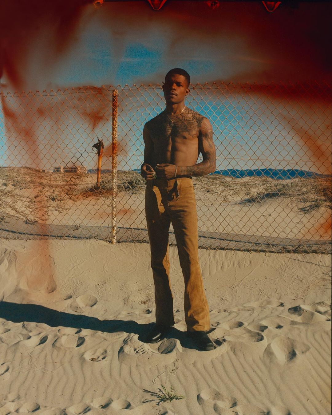 Shirtless Black man wearing tan trousers posing in sandy terrain before a chain-link fence with reddish light leaks.
