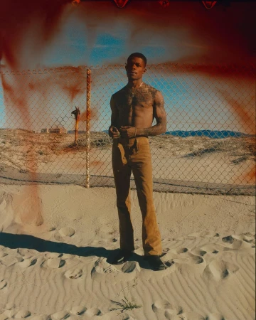 Shirtless Black man wearing tan trousers posing in sandy terrain before a chain-link fence with reddish light leaks.