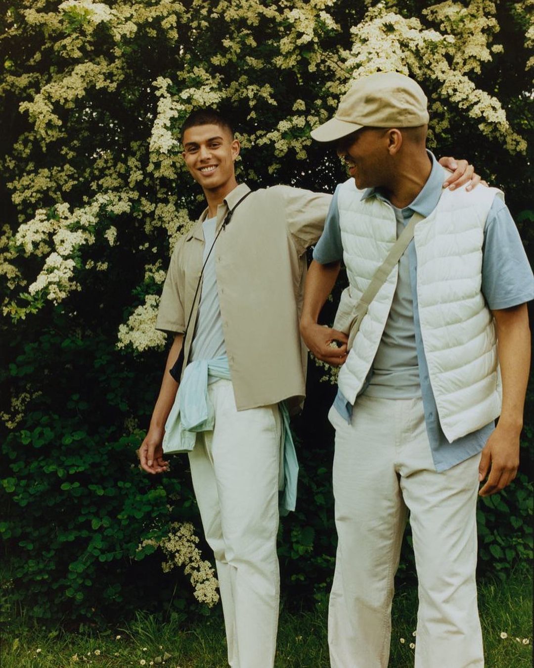 Two men in casual spring fashion, standing outdoors surrounded by flowering bushes, in natural lighting with soft color grading.