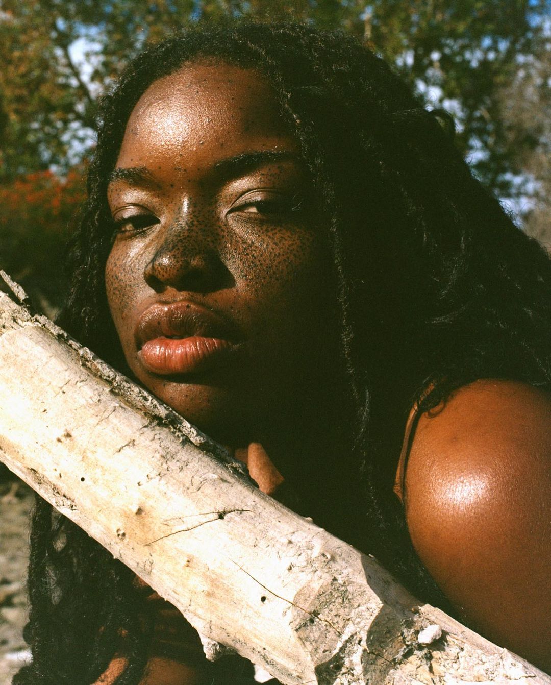 Portrait of a young Black woman with freckles and dreadlocks, leaning against a log in a natural outdoor setting with trees in the background.