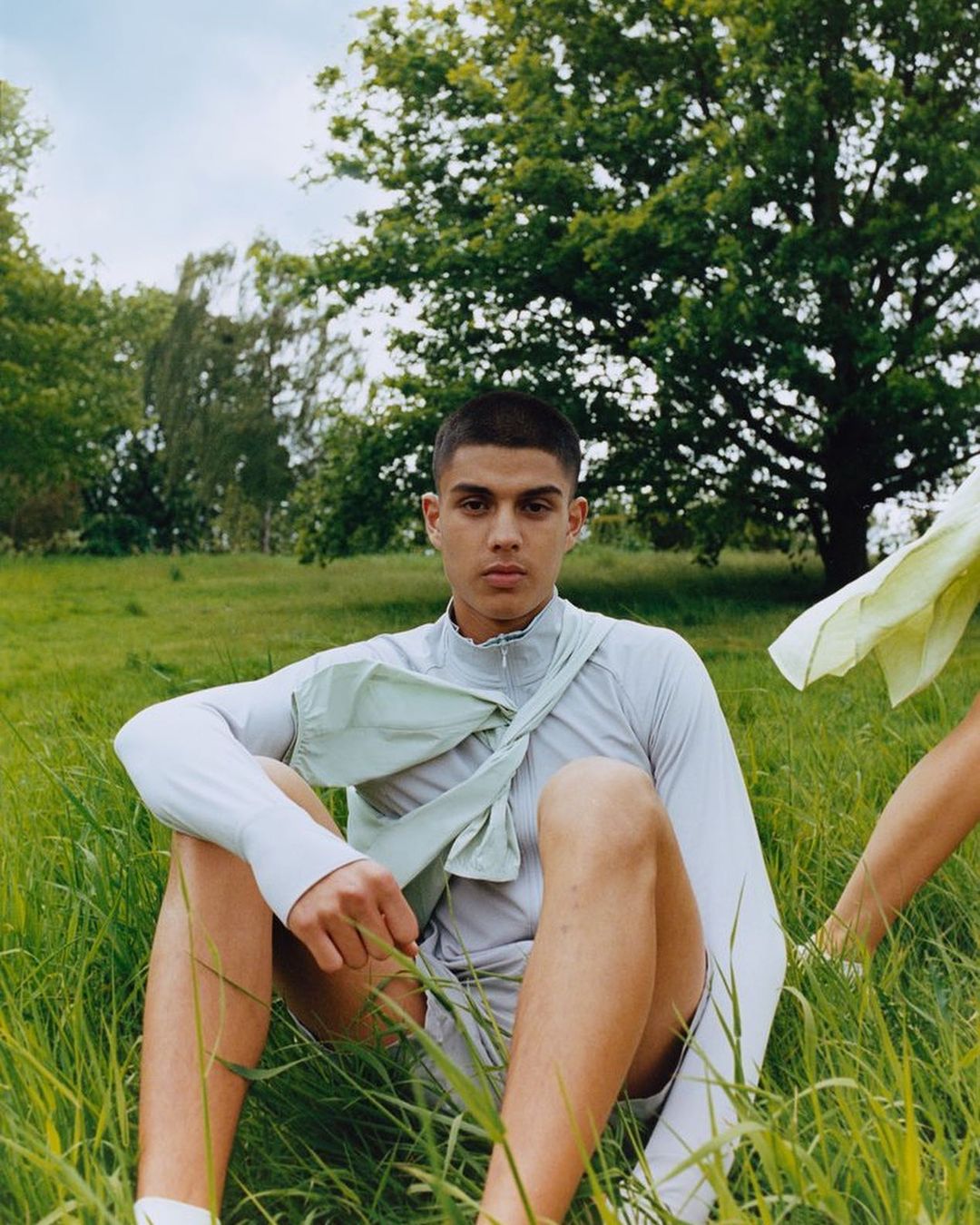 Man in a light-grey athletic outfit sitting in a grassy field with trees in the background and another person partially in the frame.
