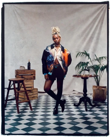 Full shot of a Black woman posing confidently in an open button-down shirt and jacket, in front of a grey backdrop with props.