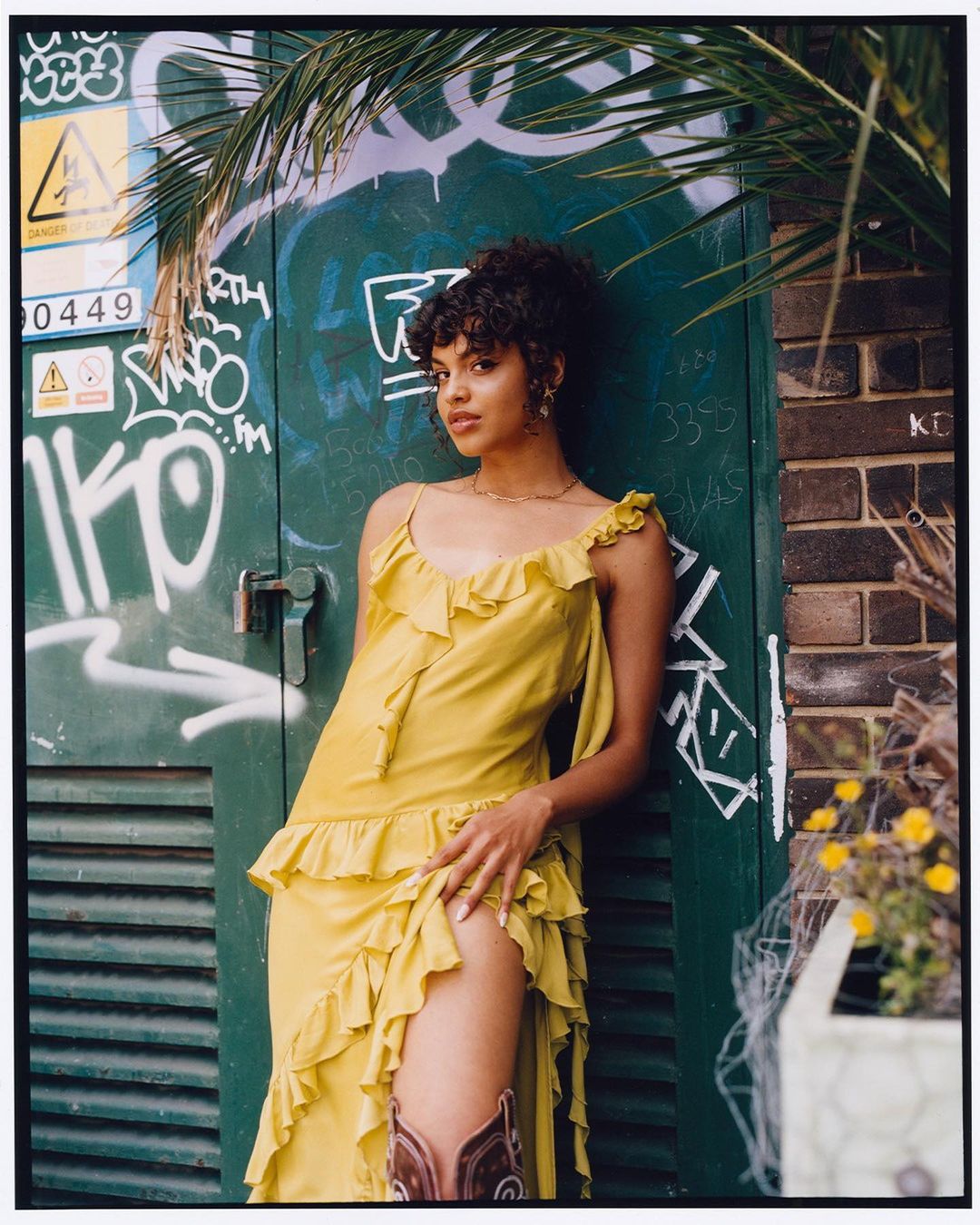 Young woman in flowing yellow dress and brown boots poses near a green door with graffiti and a palm tree overhead, outdoors.