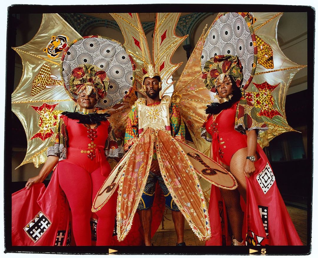 Three people in elaborate costumes with vibrant colors, ornate headdresses, and intricate details, standing indoors with backdrop.