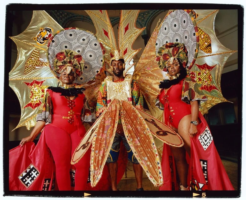 Three people in elaborate costumes with vibrant colors, ornate headdresses, and intricate details, standing indoors with backdrop.