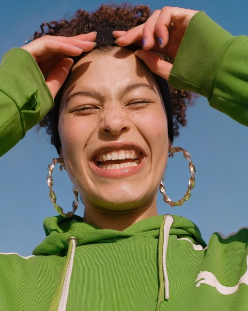A young adult woman with curly hair smiles widely in a close-up shot, wearing a green Puma hoodie and large hoop earrings against a bright blue sky.