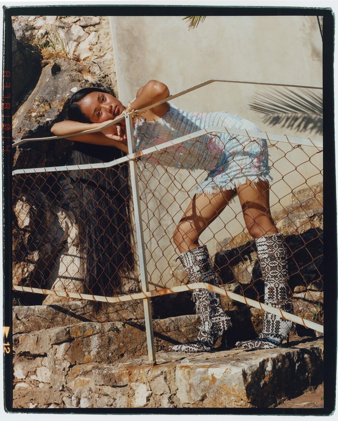 Woman in a patterned mini dress and knee-high boots leans on a rusted wire fence outside on stone steps with greenery around her.