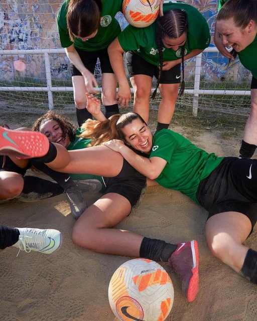 A group of happy female soccer players in green jerseys celebrate a win on a sandy pitch with a soccer ball, smiling and laughing together.
