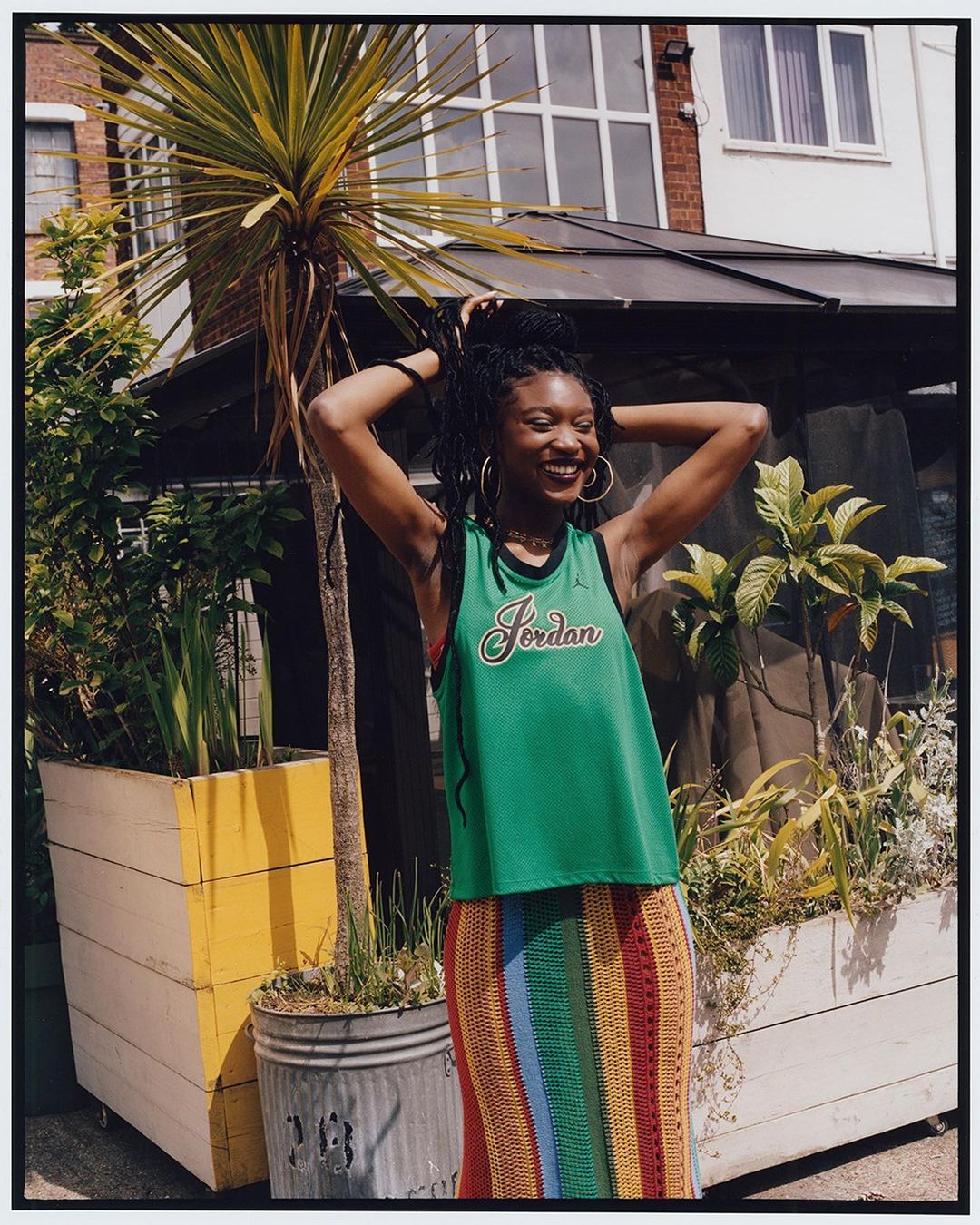 Young woman poses with hands in hair, smiling in a green 'Jordan' jersey and colorful striped skirt outdoors near plants and a house.
