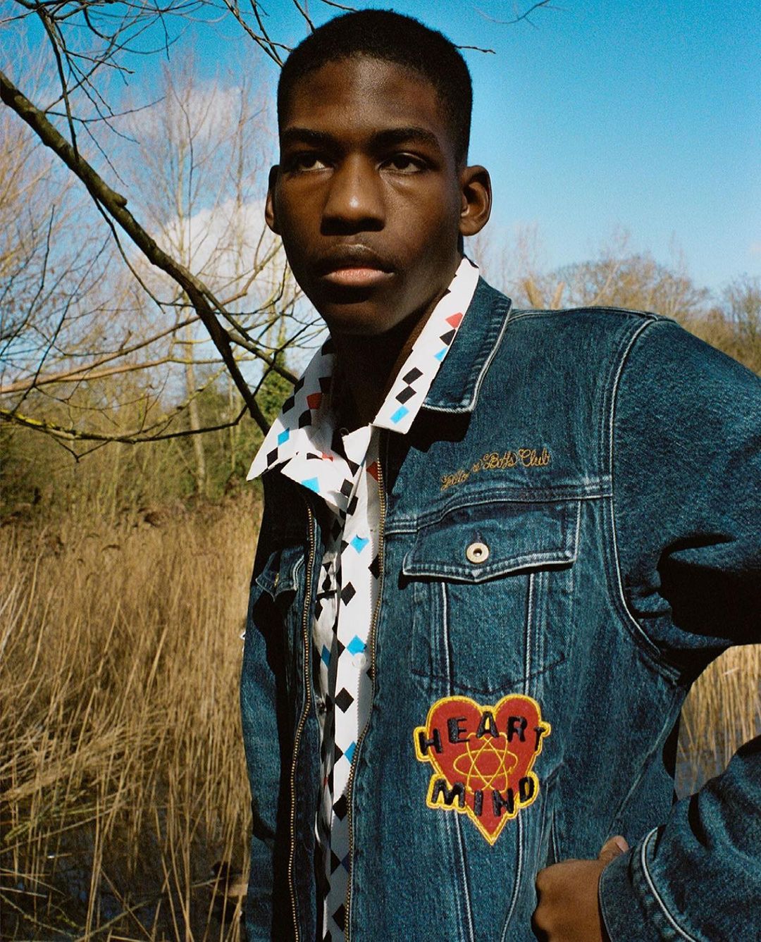 A young Black man in a denim jacket stands outdoors in a field on a sunny day, looking slightly to the left of the frame.