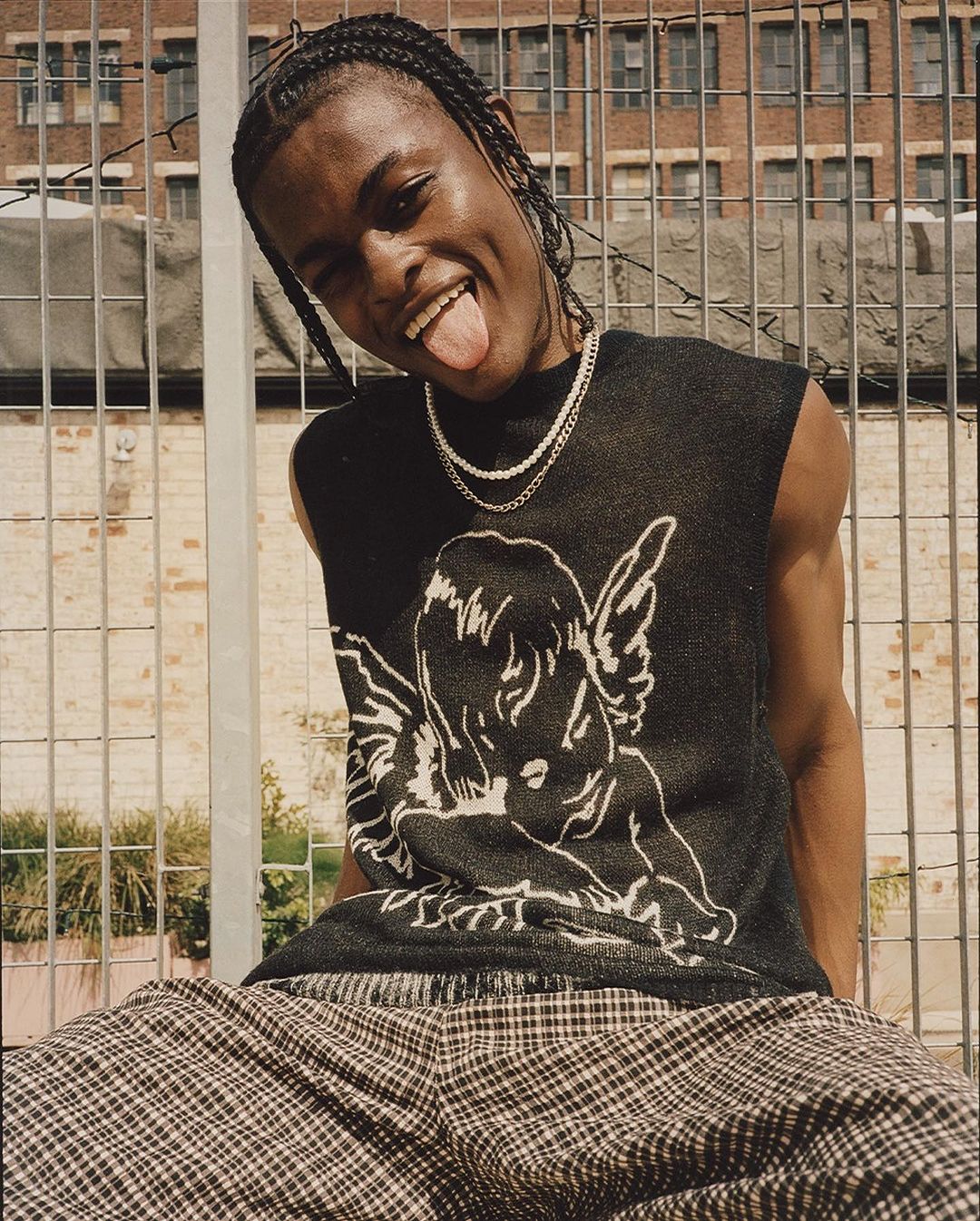 Young black man with braided hair smiling with tongue out wearing black knit vest and plaid trousers outdoors
