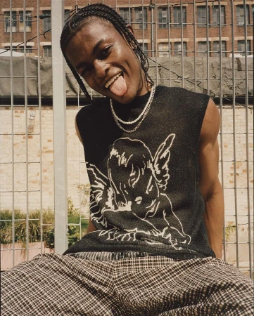 Young black man with braided hair smiling with tongue out wearing black knit vest and plaid trousers outdoors