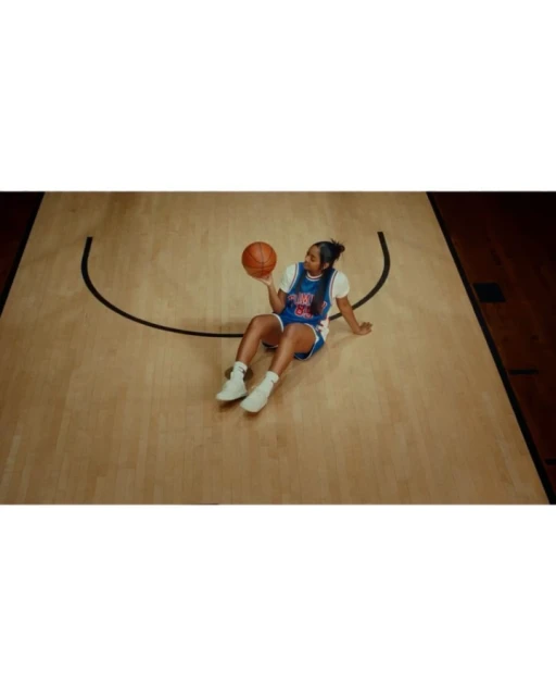 Female basketball player sitting on a court holding a basketball, wearing a blue and orange basketball jersey and white sneakers.