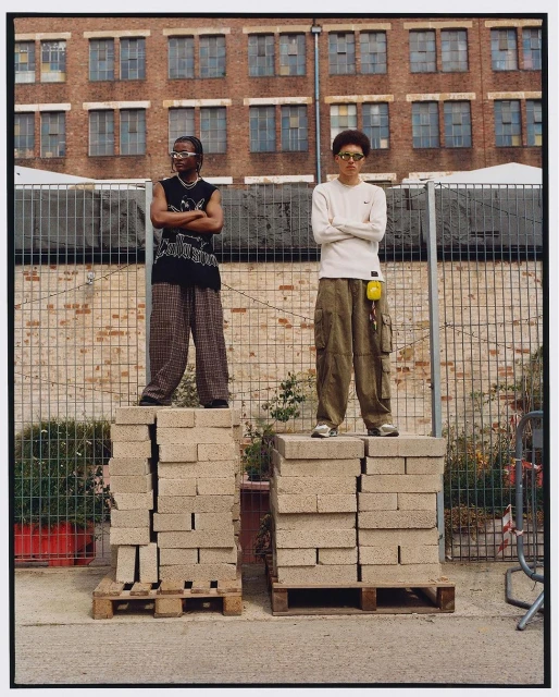 Two people stand on stacks of blocks in an outdoor urban setting, wearing casual clothes and sunglasses in front of a brick building.
