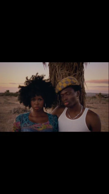 A couple posing outdoors under a tree during daytime. The woman has dark hair, and the man is wearing a patterned cap and tank top.