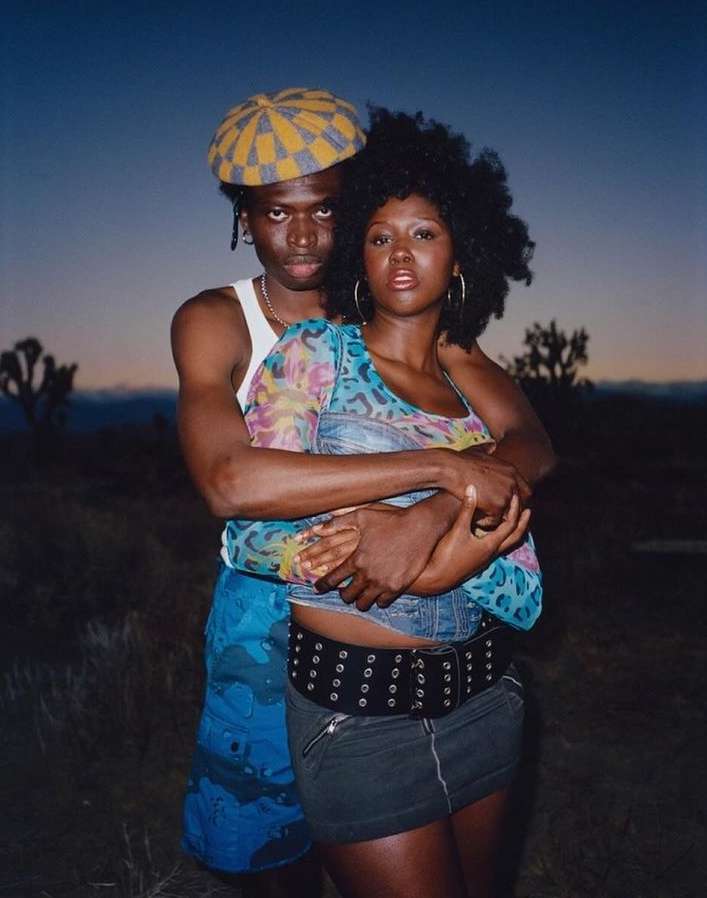 A couple embraces outdoors at dusk; the woman is in front wearing a mini-skirt and the man is behind wearing a checkered hat.