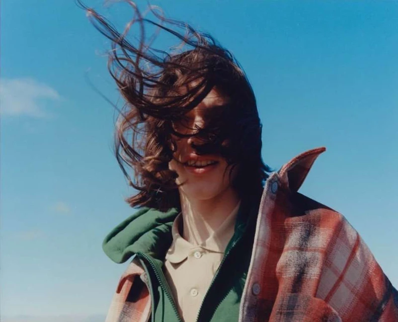 Young person with flowing hair in wind, wearing an open plaid shirt and green vest, outdoors against a blue sky on a bright day.