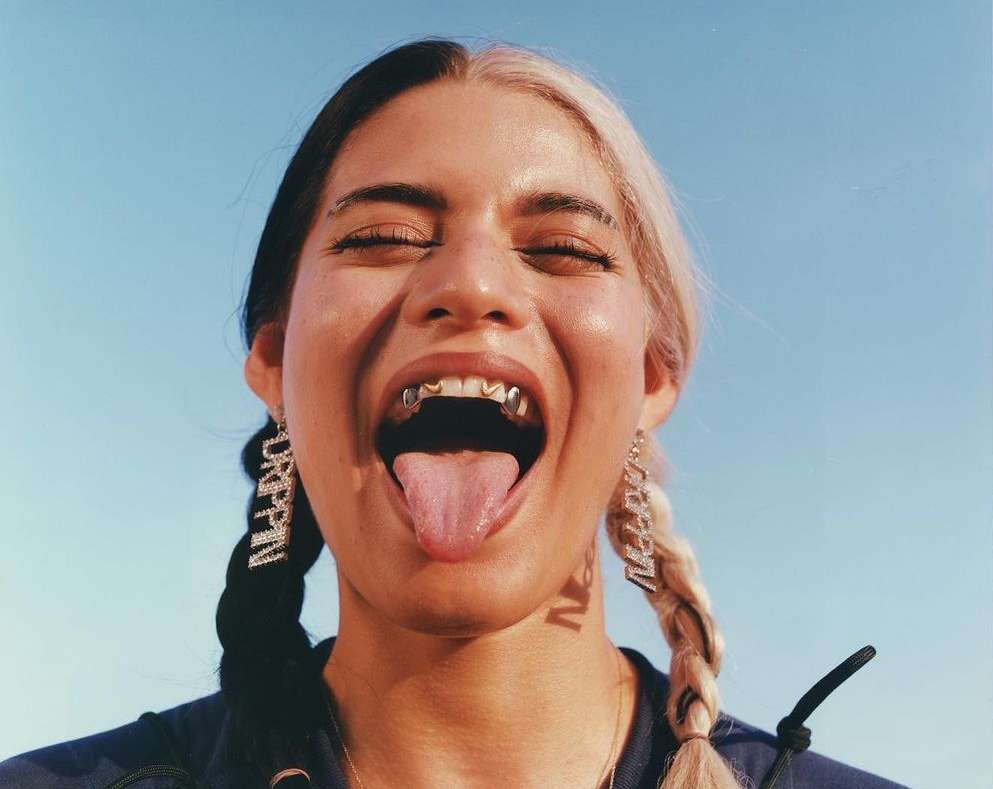 Close-up of a woman with half black and half blonde hair, gold teeth, and silver 'DRIPPIN' earrings against a clear blue sky.