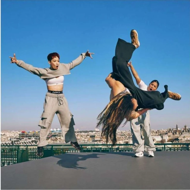 Three adult females performing a dance move on a rooftop with urban skyline background under a clear blue sky in an outdoor setting.