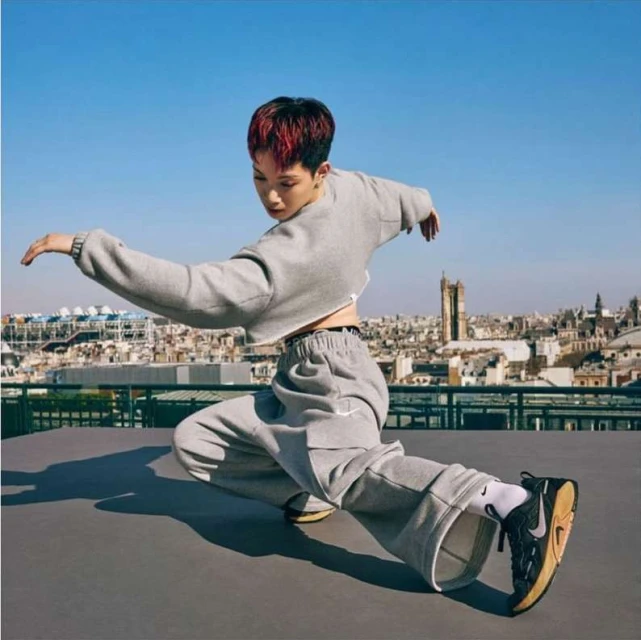 An athletic woman with a short red-dyed haircut posing in grey sweatpants and a cropped sweater on a rooftop against a cityscape backdrop.
