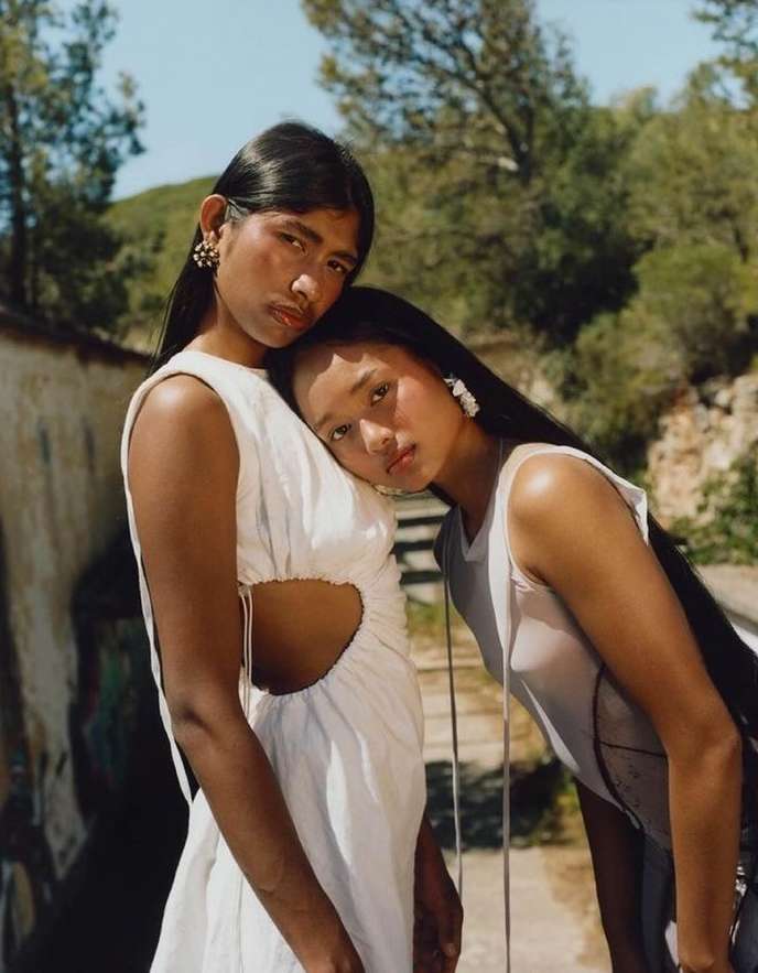 Two women pose outdoors in light-colored dresses. One woman leans head on the shoulder of the other with trees visible in background.