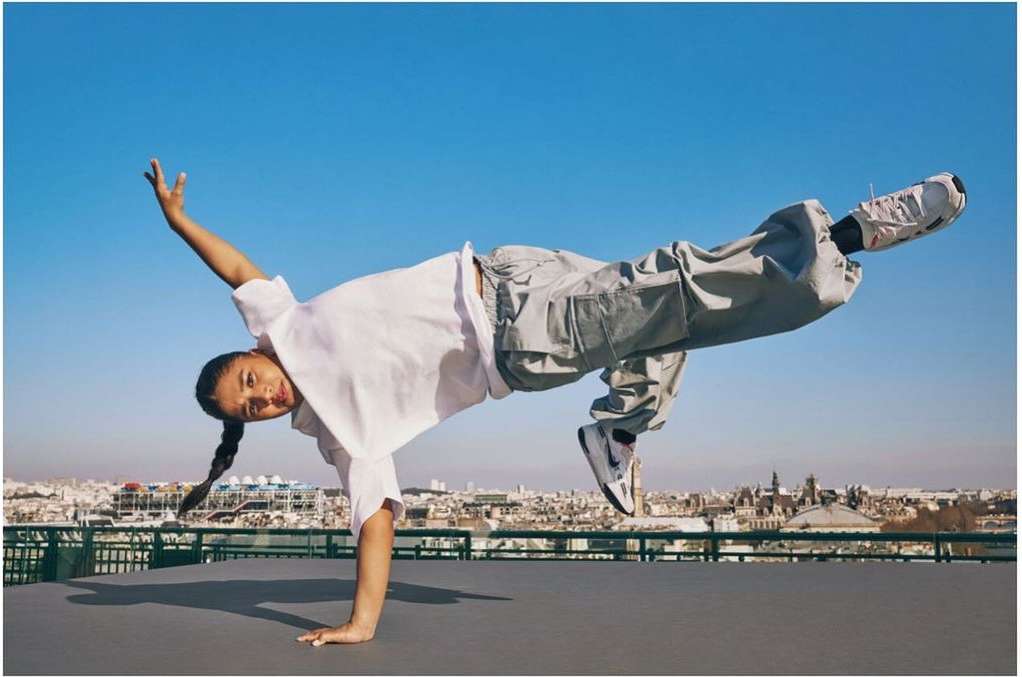 A person in dynamic pose outdoors, balancing on one hand with legs raised, wearing casual clothes and sneakers against a clear blue sky.
