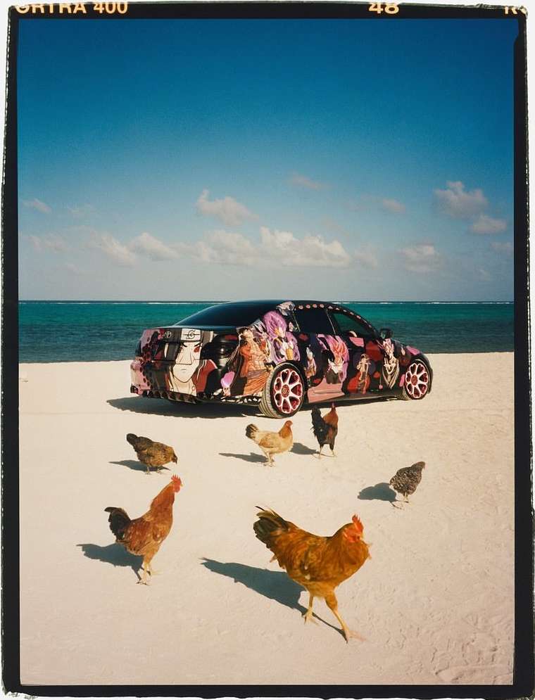 Customized car parked on a white sand beach with a group of chickens surrounding it, clear blue sky, and light turquoise ocean water.