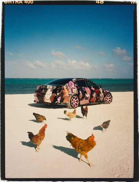 Customized car parked on a white sand beach with a group of chickens surrounding it, clear blue sky, and light turquoise ocean water.