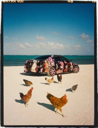 Customized car parked on a white sand beach with a group of chickens surrounding it, clear blue sky, and light turquoise ocean water.