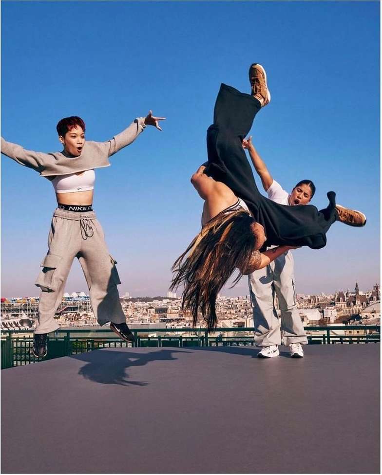 Three people in activewear pose on a rooftop with a cityscape backdrop under a blue sky. One person is being lifted upside down.
