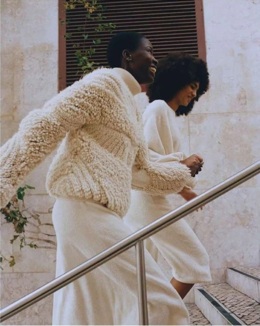 Two women walk on steps wearing casual neutral-toned clothing. One woman has short dark hair, while the other has long dark curly hair.