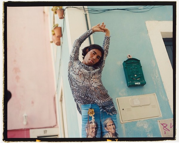 Man stretches against a light blue building on a street with pale pink buildings, showcasing artistic jeans and a patterned top in daylight.